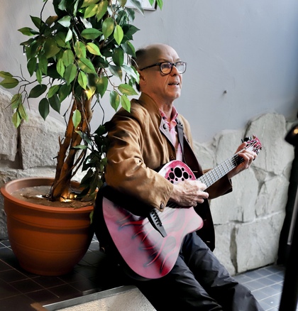 En äldre man sitter bredvid en krukväxt och spelar gitarr inomhus.