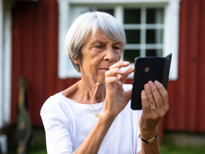 Äldre kvinna med grått hår använder en smartphone utomhus framför ett rött trähus.