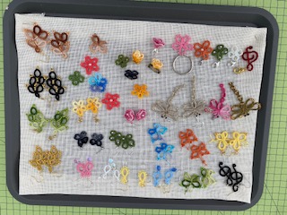 En bricka med små virkade eller knutna dekorationer i olika färger och former, placerade på ett vitt tyg.