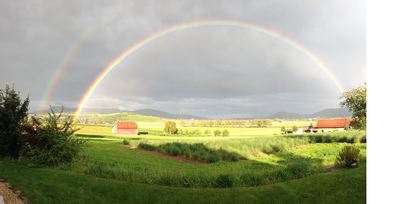 Dubbel regnbåge över ett grönt landskap med fält, hus och berg under en molnig himmel.