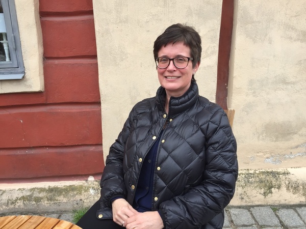 Person i svart quiltad jacka sitter vid ett runt träbord utomhus mot en putsad vägg.