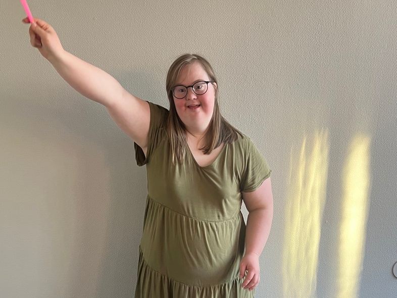 En ung kvinna med glasögon och grön klänning håller en färgglad bandstav i ett rum med ljusgrå vägg.