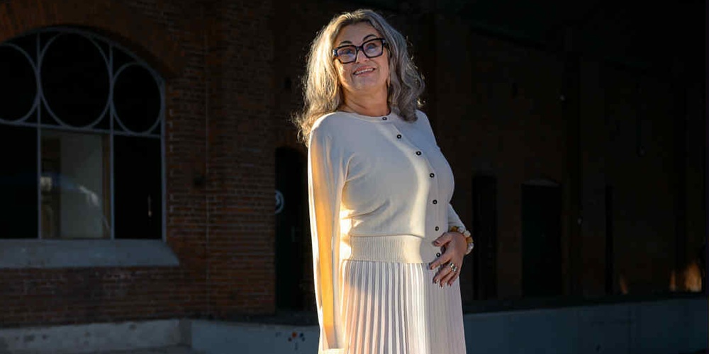 Kvinna i ljus kjol och tröja står på en perrong med tegelvägg och stora fönster i bakgrunden.