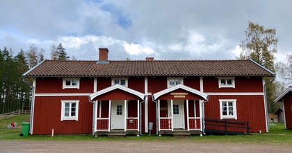 Rött trähus med vita knutar och två ingångar, omgiven av natur och träd under molnig himmel.