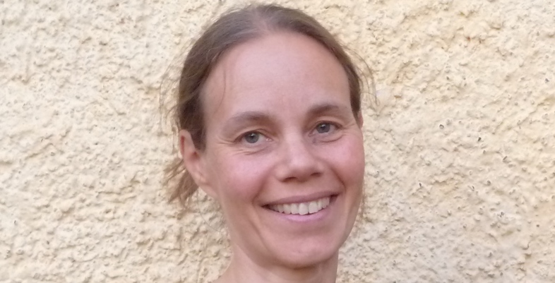 Leende kvinna med brunt hår står framför en ljusgul, skrovlig vägg.