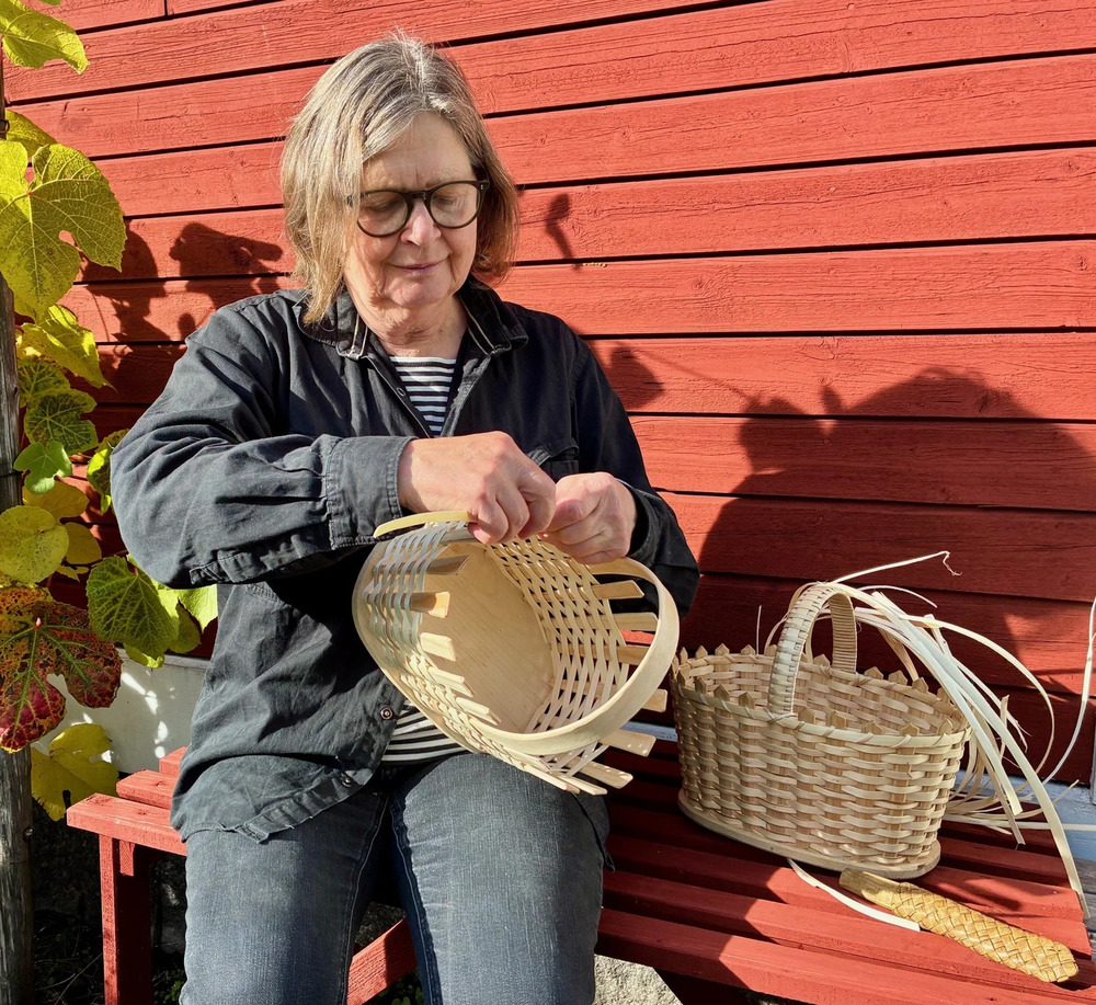 Person sitter på bänk utomhus och flätar en korg av träspån vid en röd trävägg.