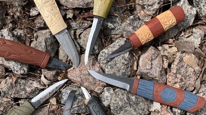Sju traditionella knivar med snidade träskaft ligger i en cirkel på stenar utomhus.