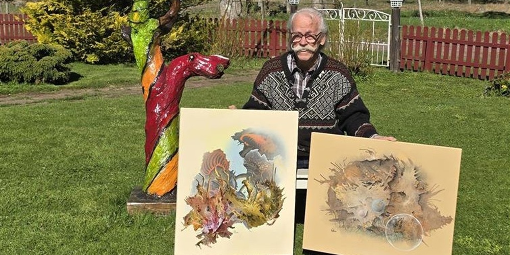 Person sitter utomhus och visar två konstverk, med en färgglad träskulptur bredvid.