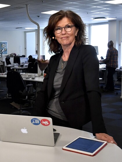 Kvinna i svart kavaj står vid ett skrivbord med en laptop och surfplatta i ett modernt kontorslandskap.