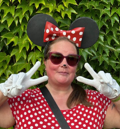 Person utklädd till Minnie Mouse med stora öron, röd prickig klänning och vita handskar, poserar framför grön växtvägg.