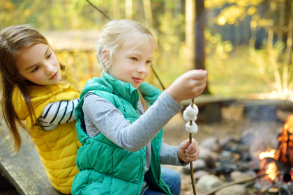 Två barn sitter vid en lägereld och grillar marshmallows på pinnar i en skogsmiljö.
