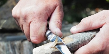 Två händer täljer en träpinne med en fällkniv utomhus.