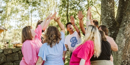 En grupp människor står i en ring utomhus och sträcker upp händerna mot mitten under ett träd.