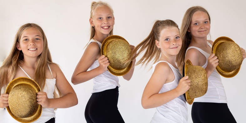 Fyra unga tjejer i vita linnen och svarta byxor poserar med glittriga guldhattar mot en ljus bakgrund.