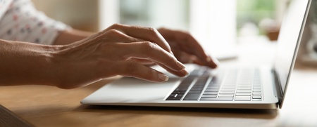 Närbild av händer som skriver på en bärbar dator vid ett träbord i ett ljust rum.