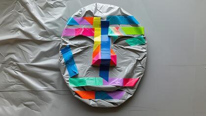 En mask täckt med silverfärgad plast och dekorerad med färgglada tejpbitar i geometriska mönster.
