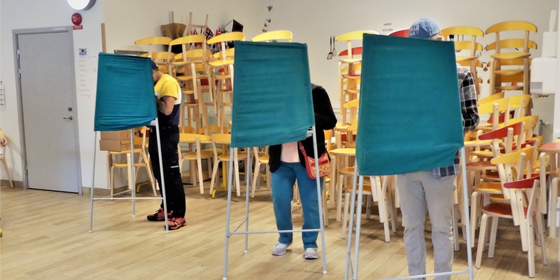 Tre personer röstar i valbås i ett rum med staplade stolar i bakgrunden.