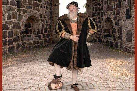 En man i historisk dräkt står på en stenbelagd gata med en avhuggen huvud vid sin fot.