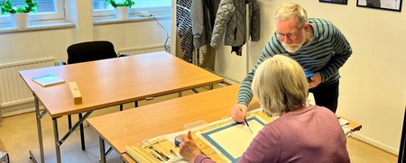 Två äldre personer arbetar tillsammans vid ett bord i ett ljust rum med konst på väggarna.