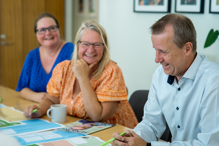 Tre personer sitter vid ett bord och skrattar under ett möte, med papper och pennor framför sig.
