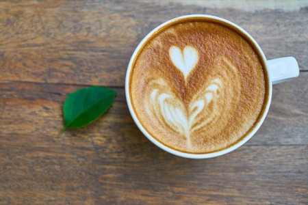 En kopp cappuccino med hjärtformad latte art står på ett träbord bredvid ett grönt blad.