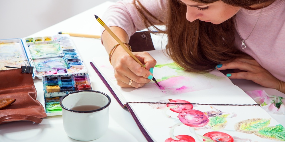 Kvinna målar akvarell i en skissbok vid ett bord med färger och en kopp te.