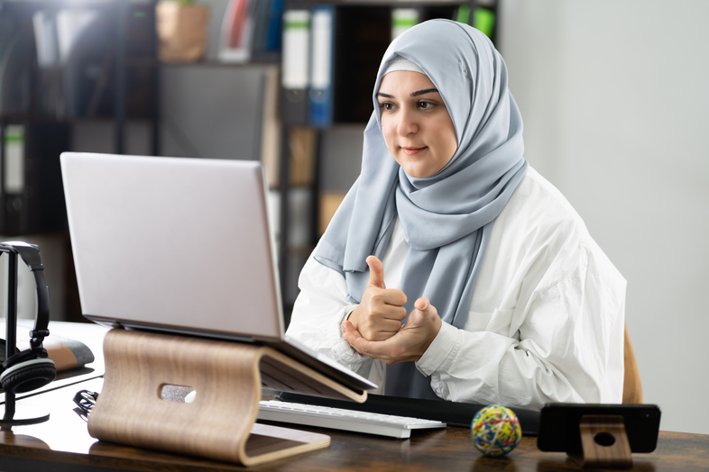 Kvinna med hijab sitter vid ett skrivbord och deltar i ett videosamtal på en bärbar dator.