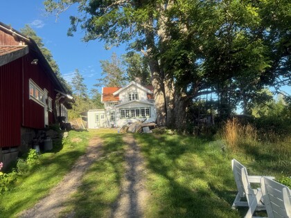En grusväg leder fram till ett vitt hus med rött tak, omgiven av träd och gräs under en solig dag.