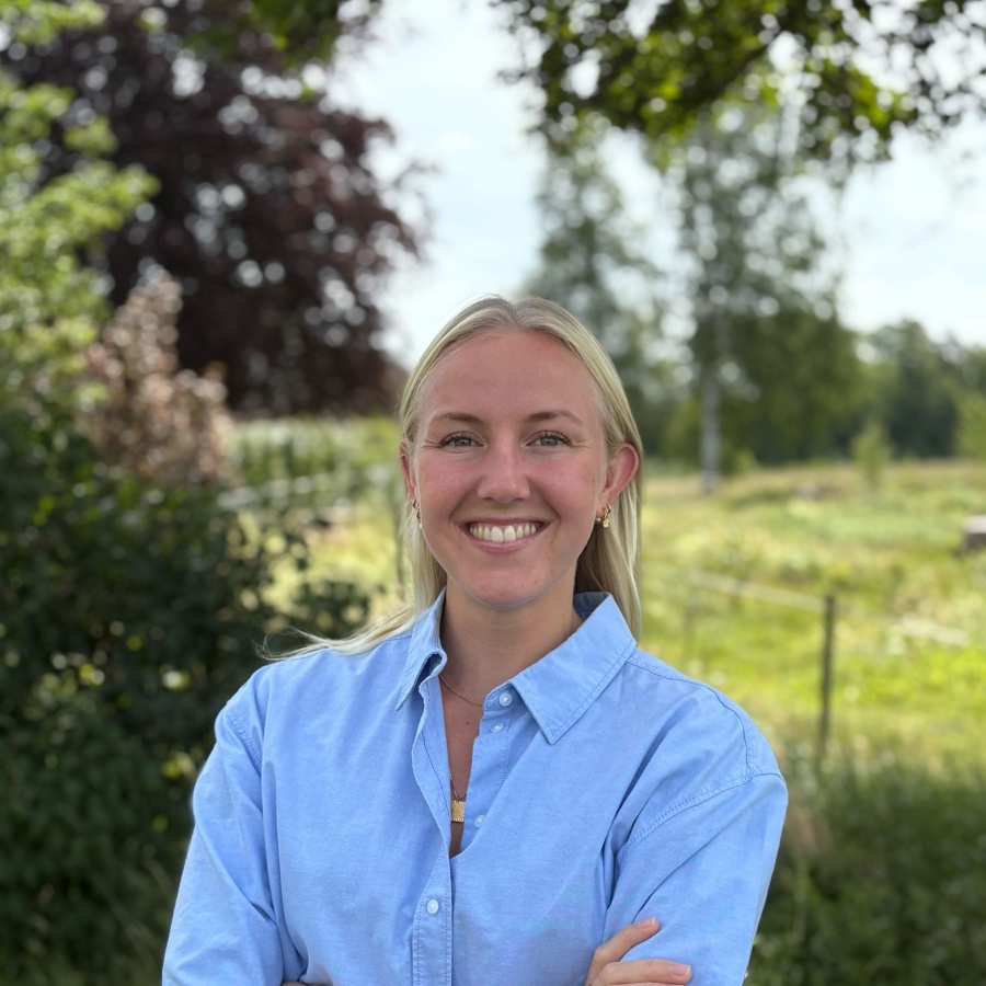 Leende kvinna med blont hår i blå skjorta står utomhus med armarna i kors.