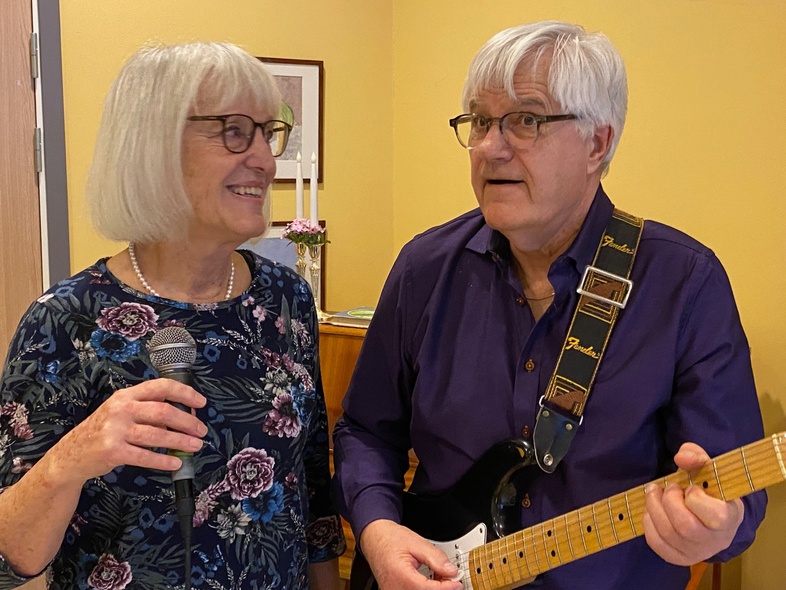 En äldre kvinna sjunger i mikrofon och en äldre man spelar elgitarr i ett rum med gul vägg.