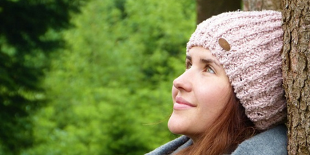 Person med stickad mössa och halsduk lutar mot ett träd i en grön skogsmiljö.