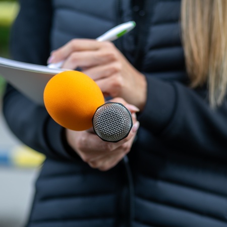 En person håller en mikrofon med orange skydd och antecknar i ett block utomhus.