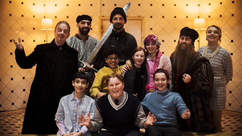 En grupp människor i färgglada och varierande kläder poserar tillsammans inomhus mot en mönstrad vägg.