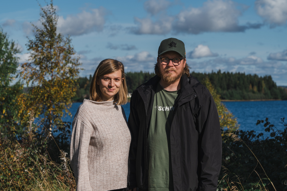 En kvinna och en man står utomhus vid en sjö med skog i bakgrunden under en solig dag.