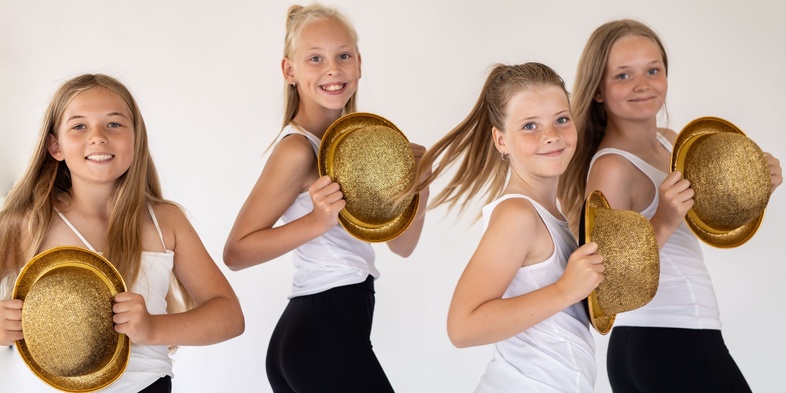 Fyra unga tjejer i vita linnen och svarta byxor poserar med glittriga guldhattar mot en ljus bakgrund.