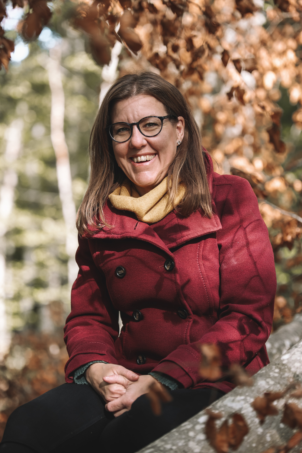 Kvinna med glasögon och röd kappa sitter utomhus bland höstlöv och ler mot kameran.