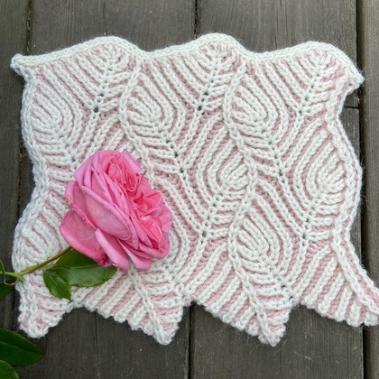 Stickad bit i vitt och rosa med ett lövmönster, dekorerad med en rosa ros ovanpå, placerad på träbord.