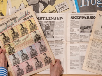 Flera gamla tidningar och programblad på svenska ligger utspridda på ett bord, en person bläddrar bland dem.