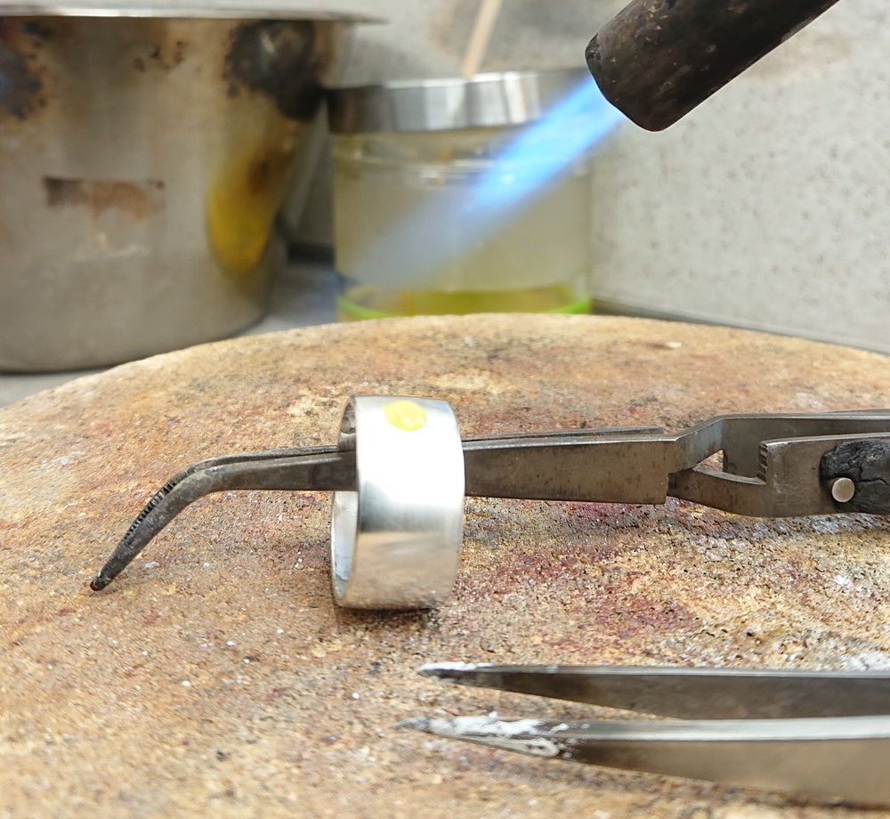 En ring värms upp med en blå gaslåga, hålls med en tång på en arbetsyta i ett smyckesverkstad.