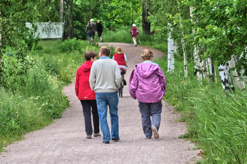Flera personer promenerar på en grusväg omgiven av gröna träd och gräs en sommardag.