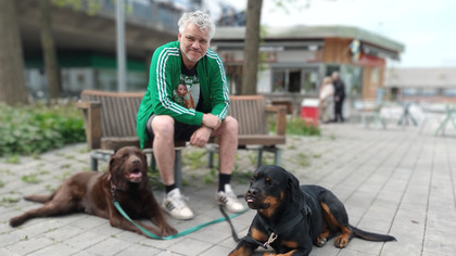 En man sitter på en bänk utomhus med två hundar, en brun och en svart, liggande framför sig.