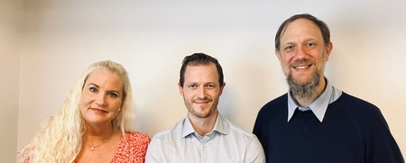 Tre personer står bredvid varandra mot en ljus vägg i ett kontorsliknande rum.