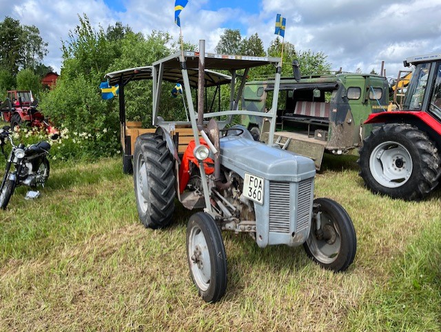 En äldre grå traktor med svensk flagga står parkerad på en gräsmatta bland andra fordon.