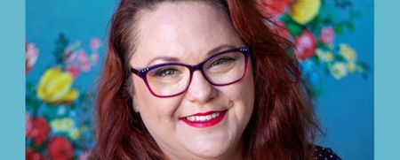Leende kvinna med långt rött hår och glasögon framför en färgglad blommig bakgrund.