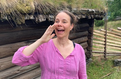 Person i rosa skjorta står utomhus vid en gammal träbyggnad och håller handen vid huvudet.