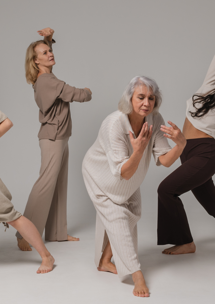 Fyra kvinnor i olika åldrar dansar barfota i mjuka, neutrala kläder mot en ljus bakgrund.