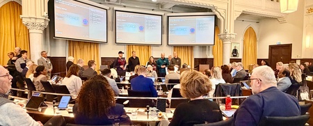 Möte i en stor sal med många personer sittande runt ett bord och presentation på skärmar.