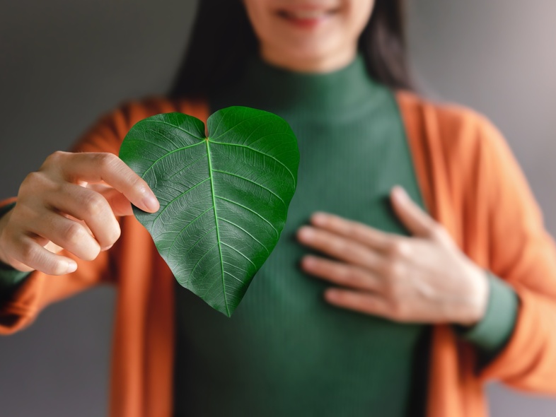 Person håller ett grönt hjärtformat blad framför bröstet, klädd i grön tröja och orange kofta.