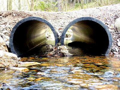 Två stora rör under en grusväg med vatten som rinner igenom, omgivna av stenar och natur.