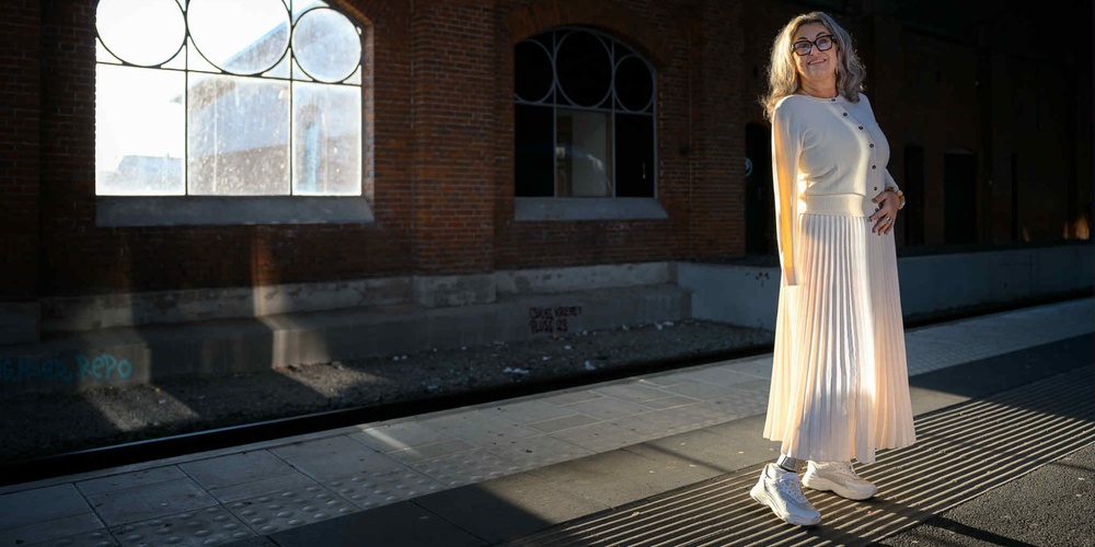 Kvinna i ljus kjol och tröja står på en perrong med tegelvägg och stora fönster i bakgrunden.